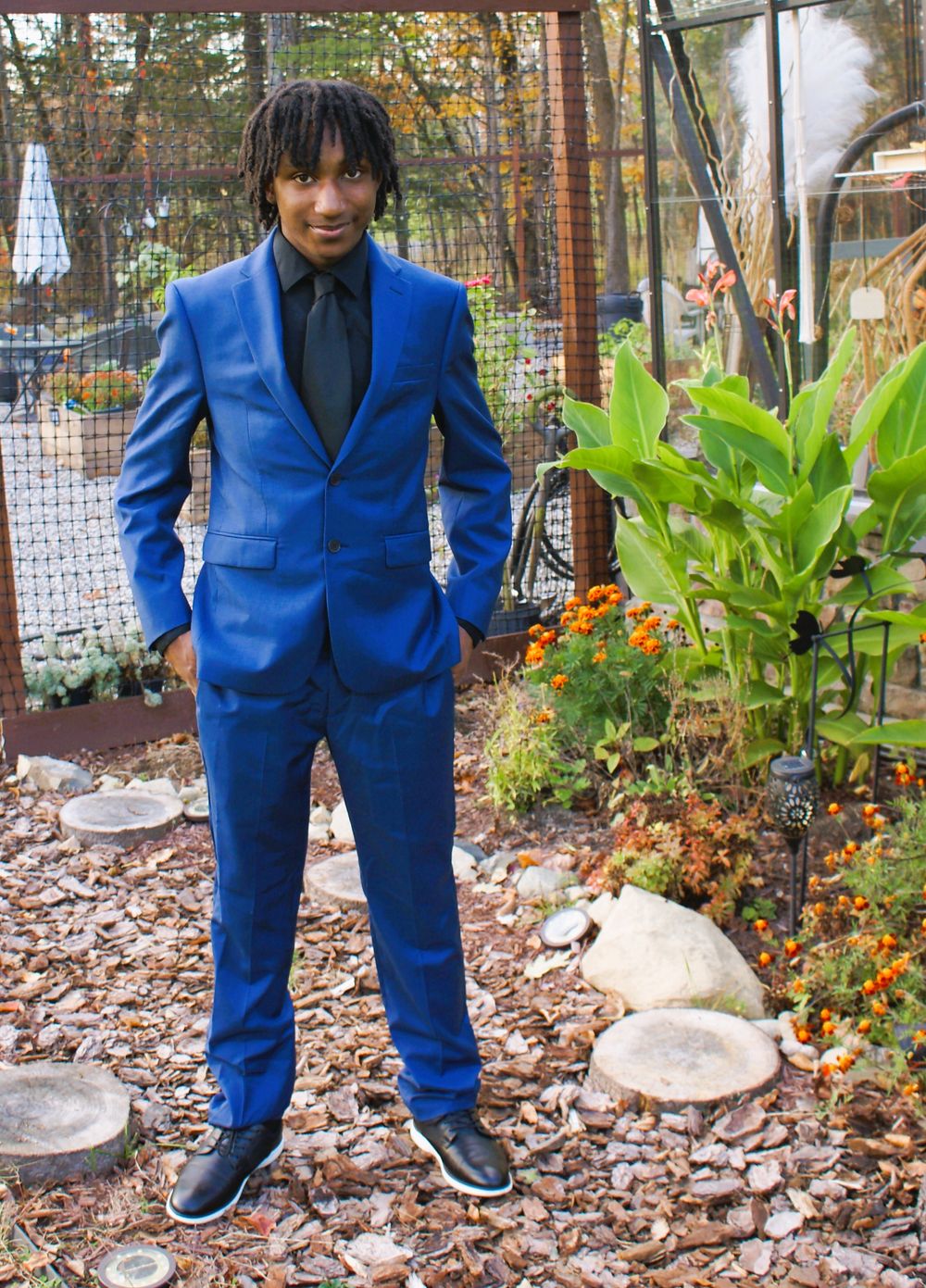 Young man in a bright blue suit with black shirt and tie posing outdoors in a garden setting.