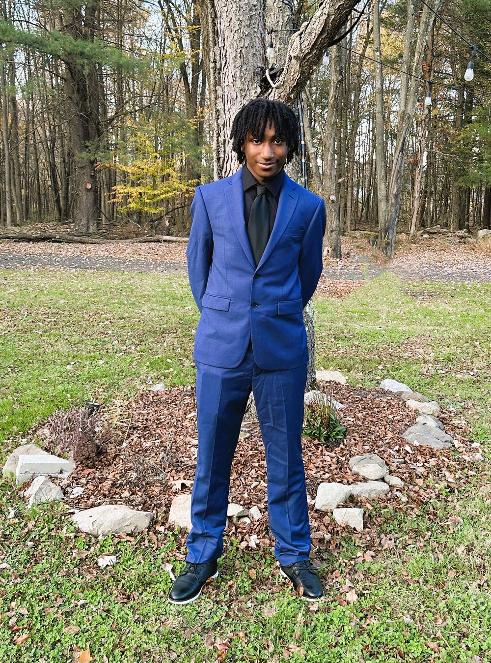 Young man in a blue suit and black shirt posing outdoors in a wooded area with fall foliage.
