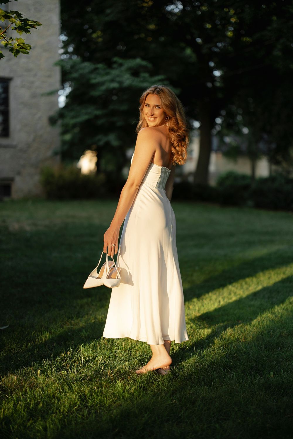 Radiant bride in a white halter dress walks barefoot on grass, holding heels at golden hour.