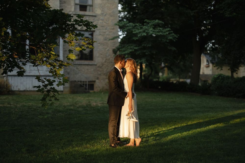 Newlyweds share a tender kiss on a sunlit lawn with a historic building in the background.