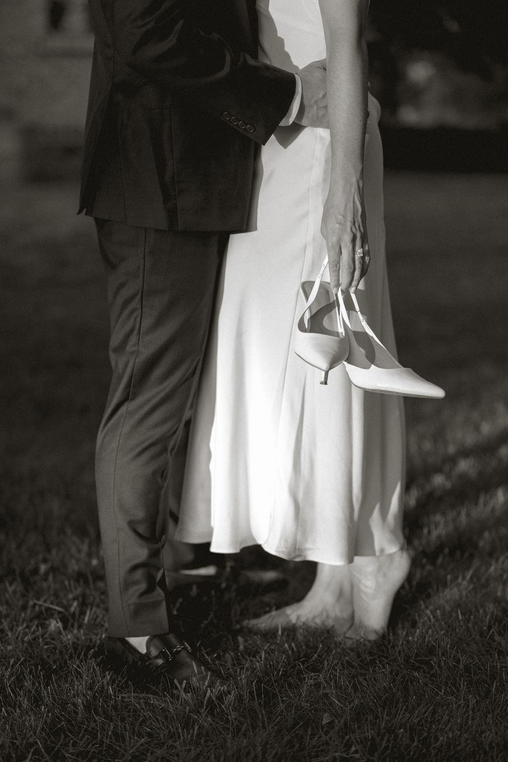 Black and white photo of bride and groom embracing on grass, bride holding white heels.