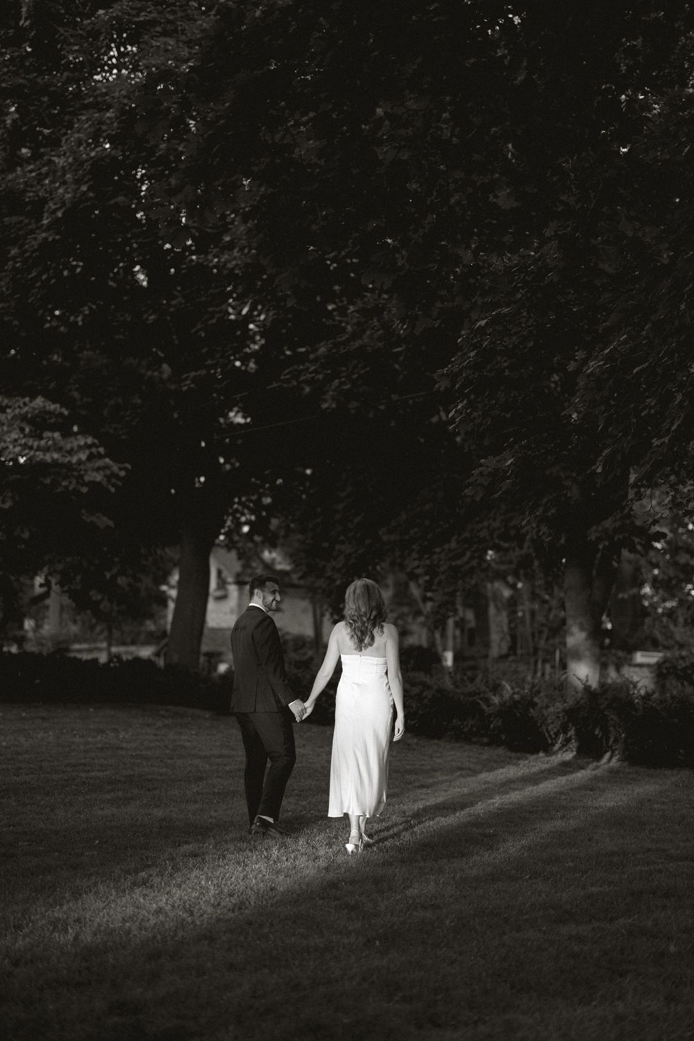 Black and white image of newlyweds walking hand in hand through a tree-lined garden path.