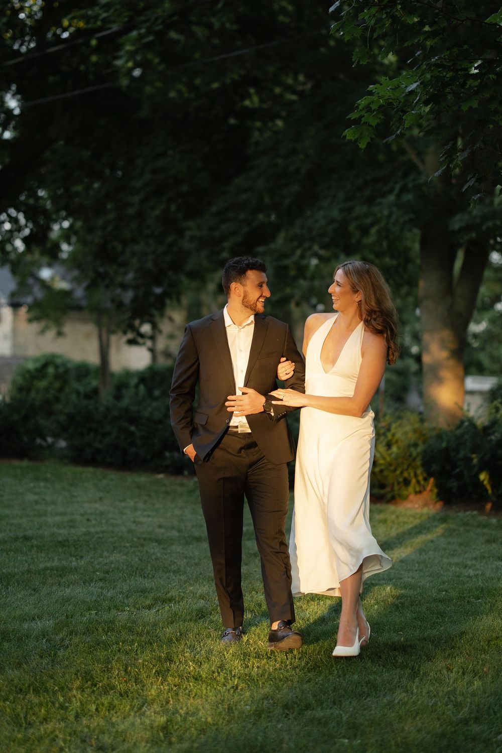 Smiling couple in wedding attire walk together on a lush green lawn at golden hour.