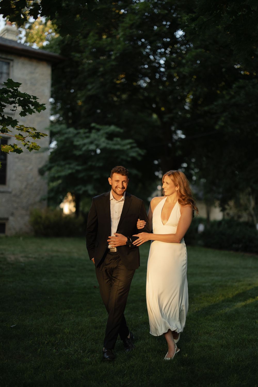 Elegant couple strolling arm in arm on a sunlit lawn beside a charming stone building.