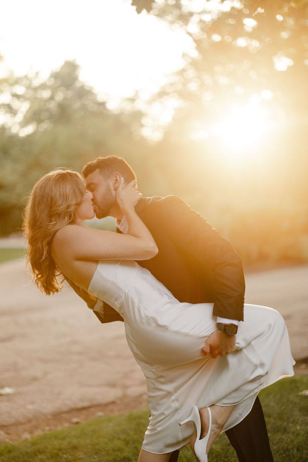 Groom lifts bride in a passionate kiss bathed in stunning golden sunset light outdoors.