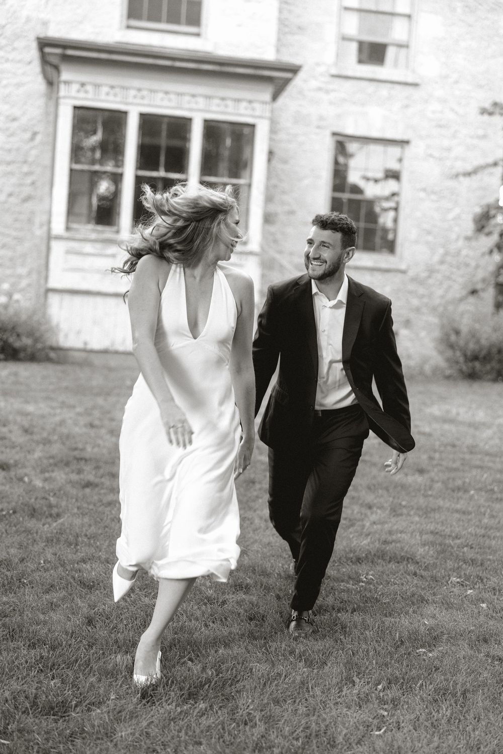 Black and white photo of smiling bride and groom running across lawn outside historic stone building.