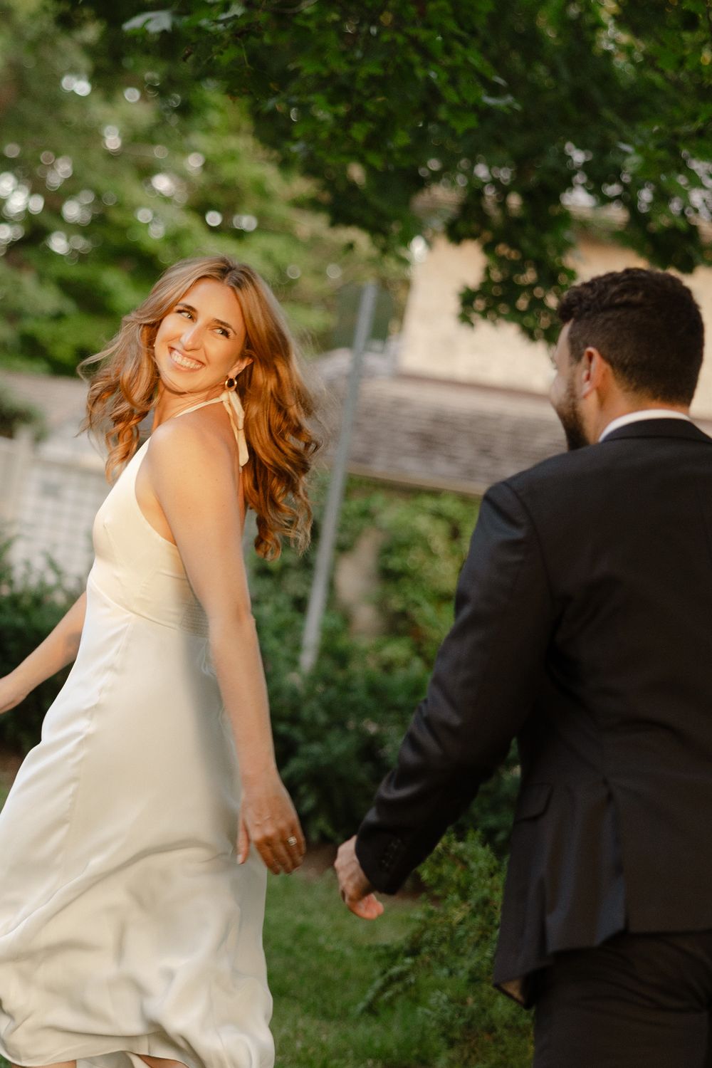 Bride in white halter gown smiles back at groom while holding hands in lush garden at golden hour.