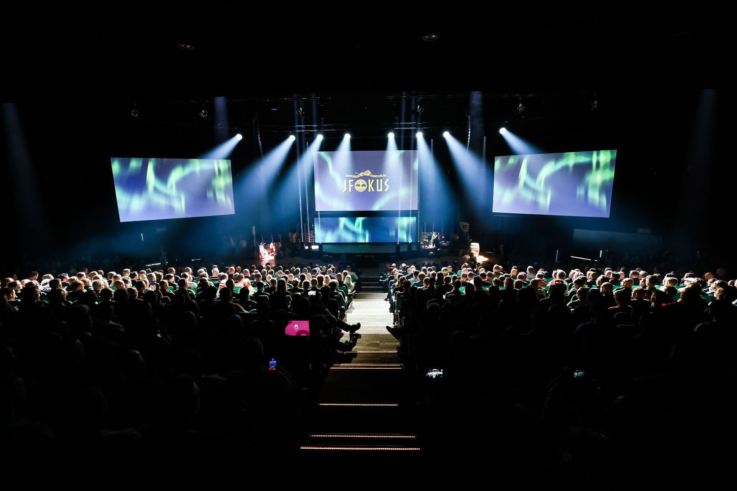 Large orchestra performing on a grand stage with green aurora borealis screen displays.