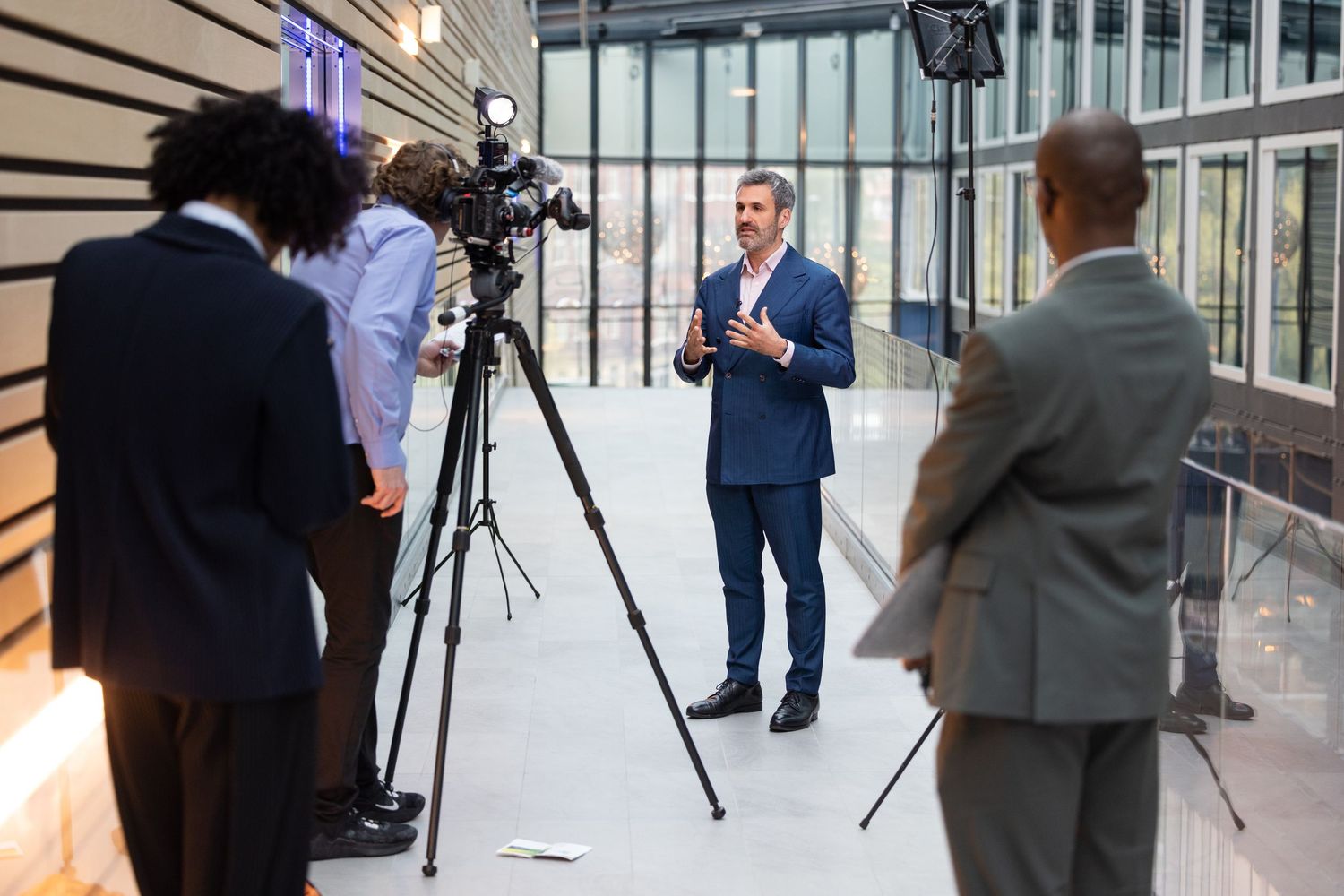 Businessman in blue suit being filmed by camera crew in a modern glass building atrium.