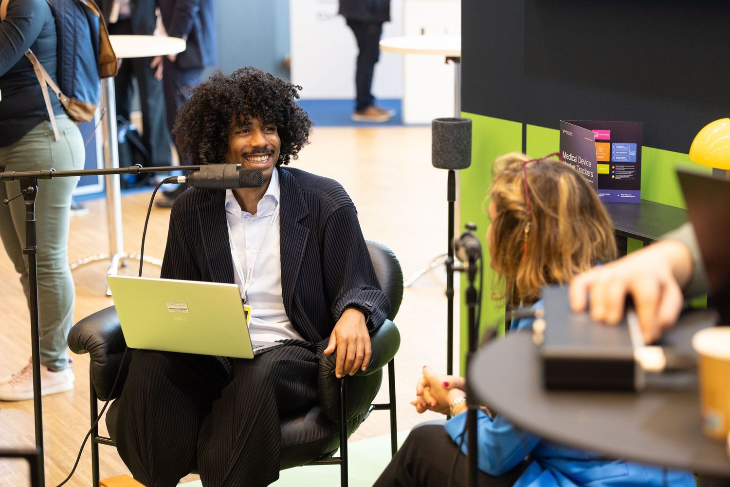 Smiling man with curly hair using laptop seated at a tech conference networking area.
