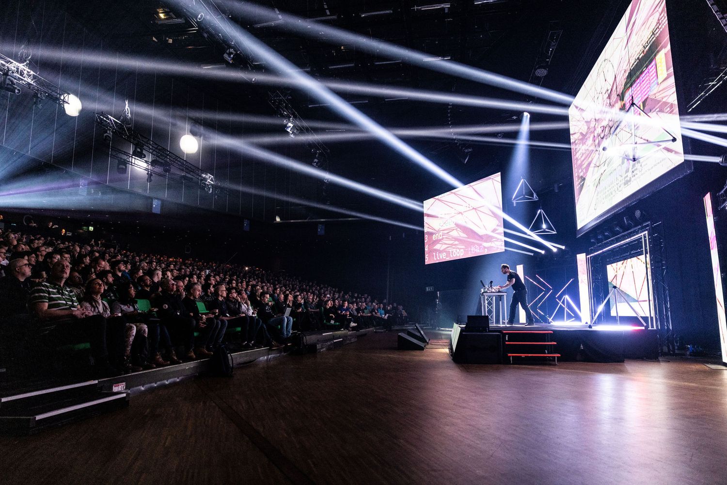A speaker presents on stage before a large audience in a dark concert venue.