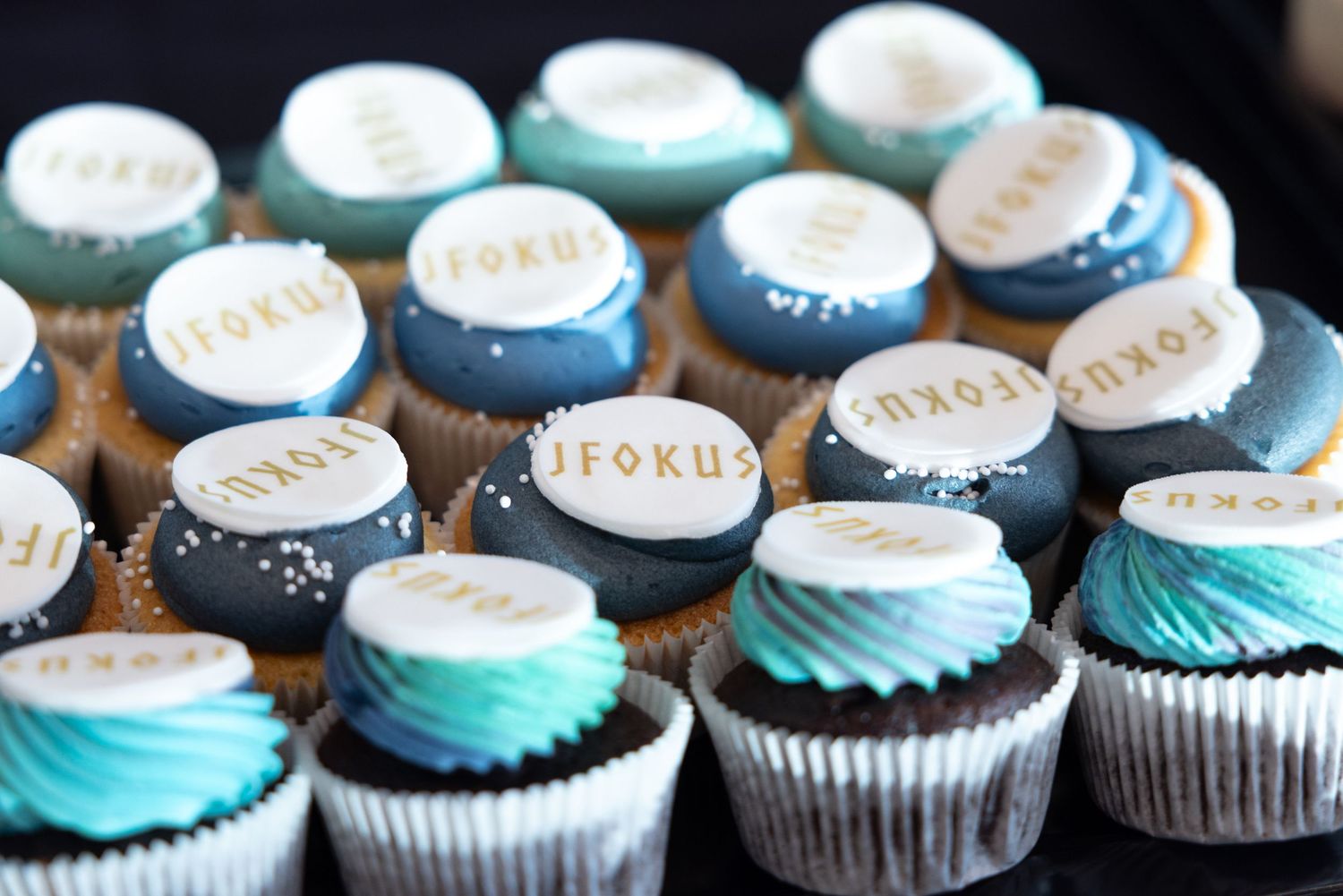 Decorative cupcakes with teal and white frosting topped with round fondant discs reading FOKUS at a tech event.