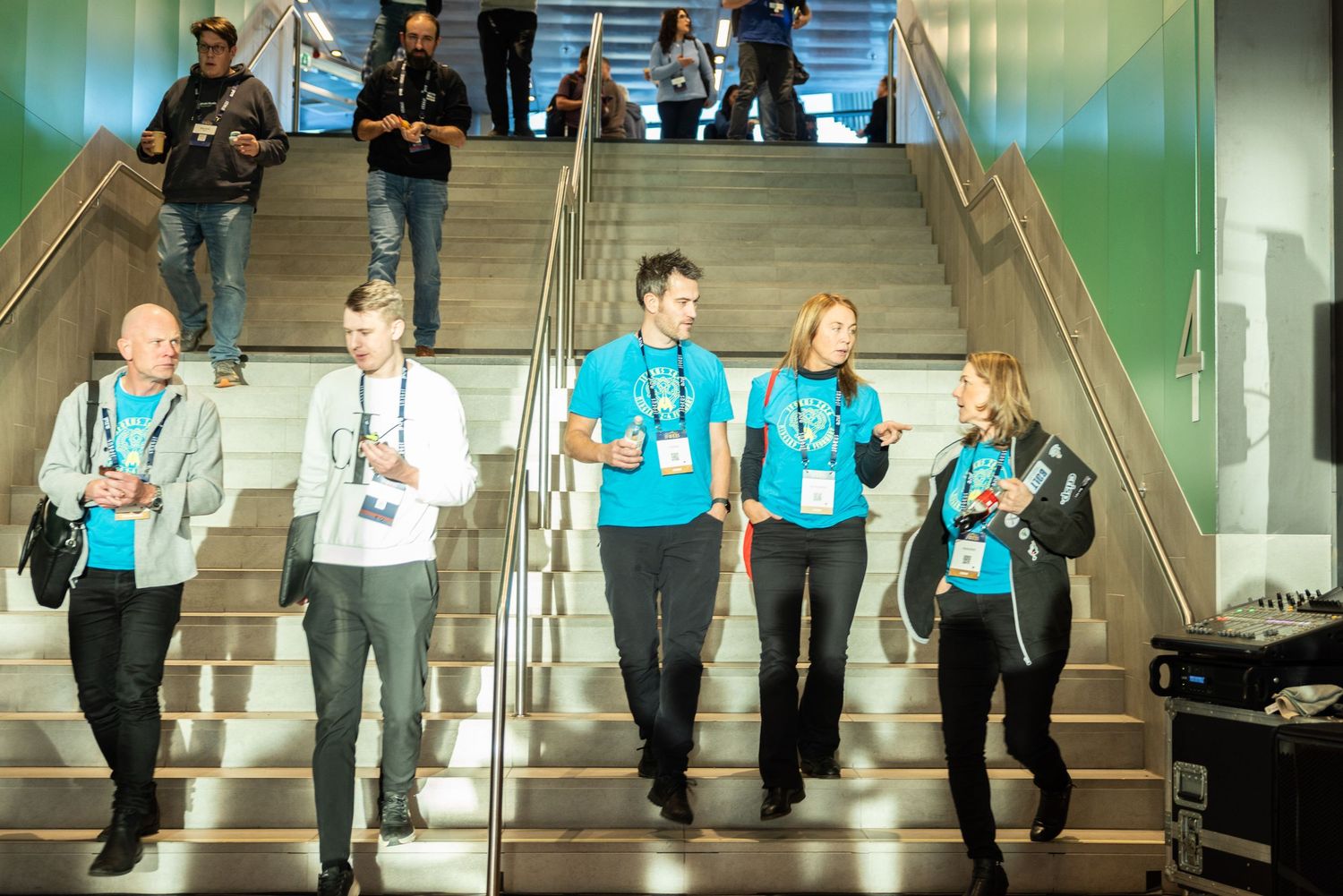 Conference staff wearing teal lanyards walking down modern stadium stairs at a tech event.