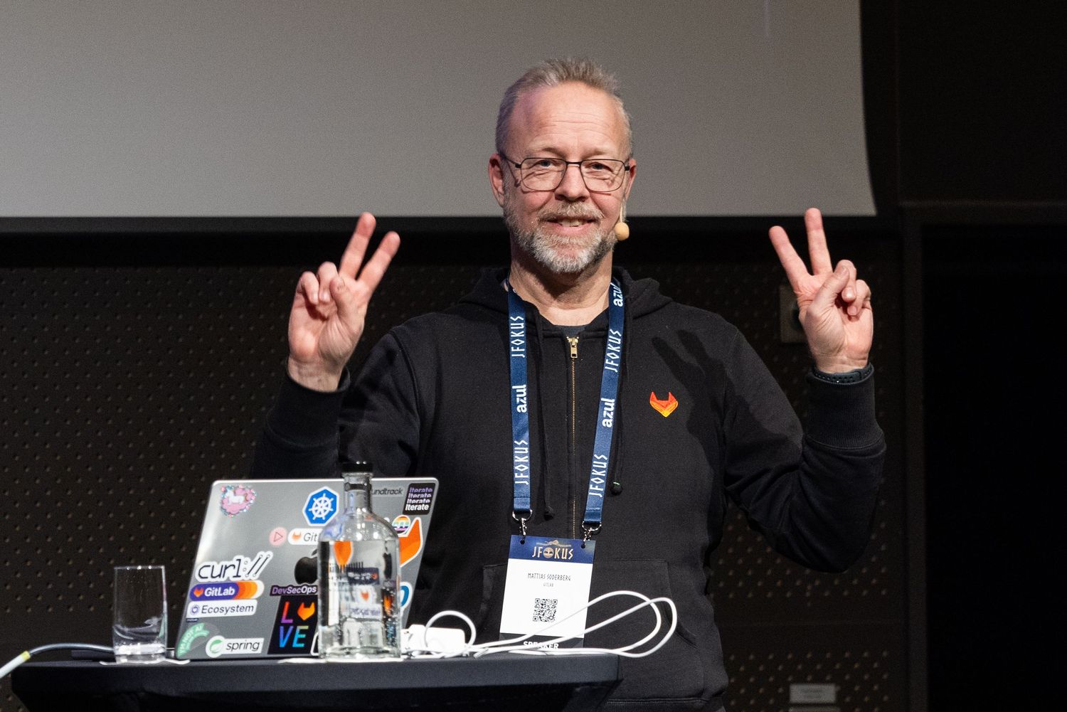 Smiling male speaker making peace signs at a tech conference presentation desk with laptop.