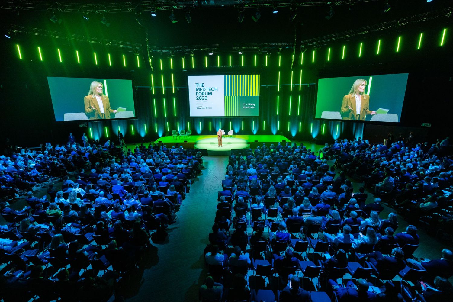 Large conference auditorium filled with attendees watching a speaker on a lit stage.