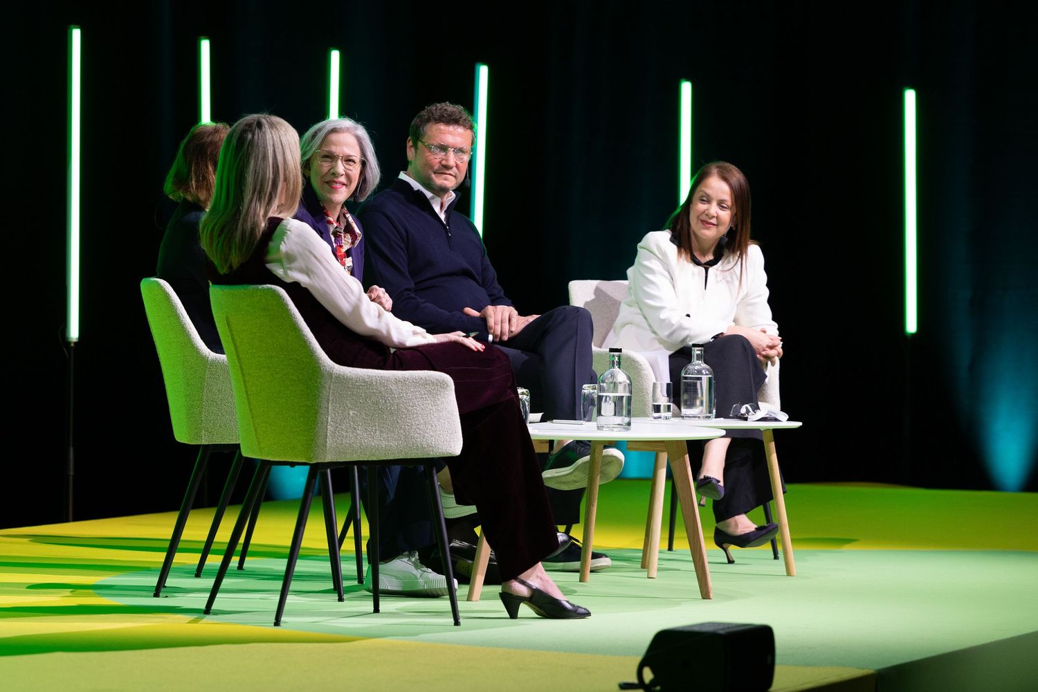 Four panelists engaged in discussion on a green-lit stage at a professional conference event.