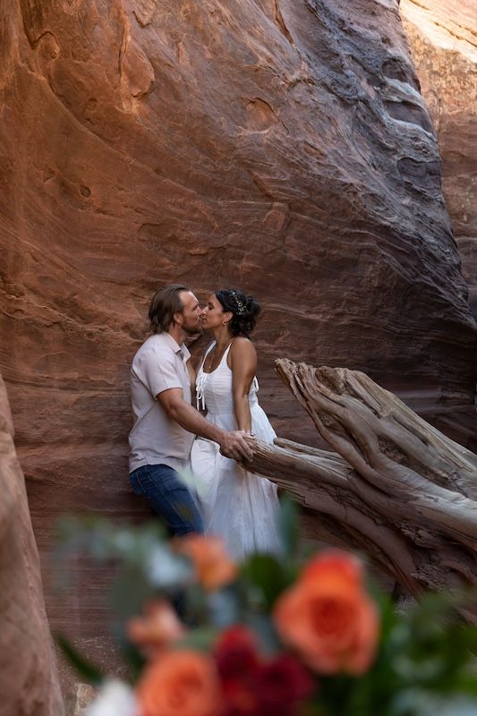 Couple sharing a romantic kiss in a stunning red rock canyon, surrounded by driftwood and colorful roses in the foreground.