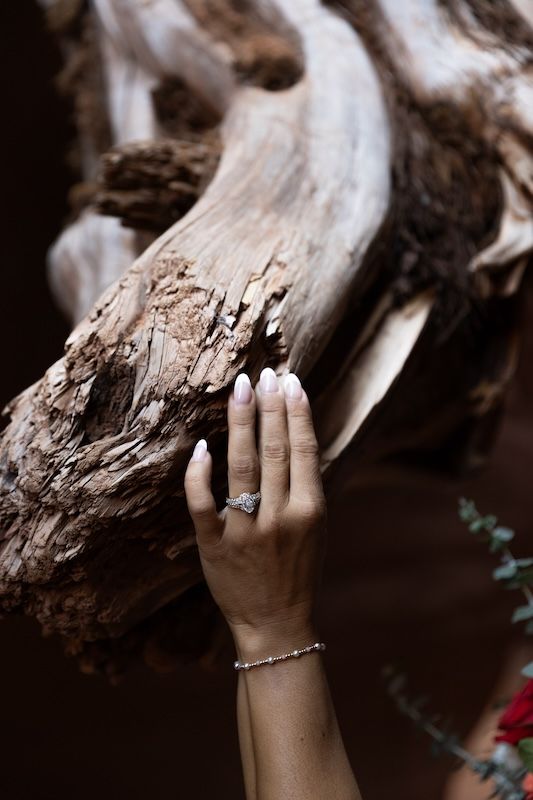 A woman's hand with white nails and a diamond ring rests on weathered driftwood, wearing a delicate silver bracelet.