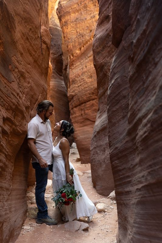 Couple sharing a romantic moment in a stunning red rock slot canyon, bride holding red roses in a white dress.