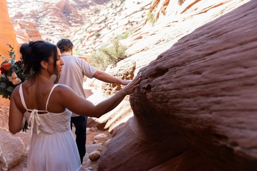 Couple in wedding attire touching red sandstone rock formation in a desert canyon setting.
