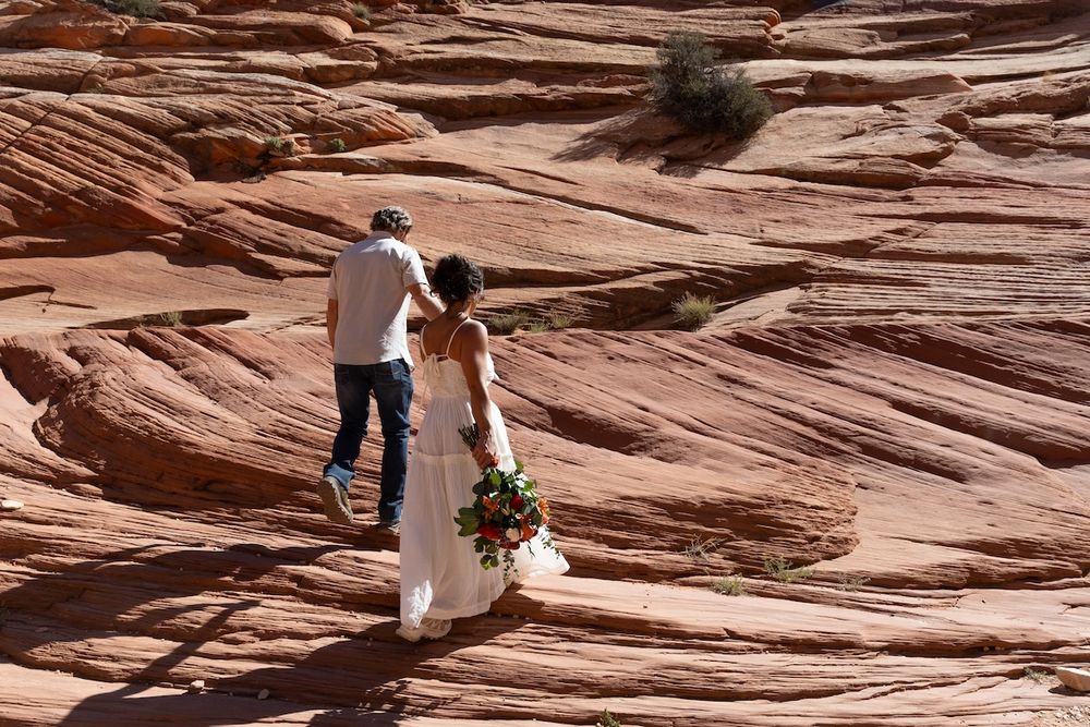 Bride in white gown holding bouquet with groom on red rock sandstone formations in desert landscape.