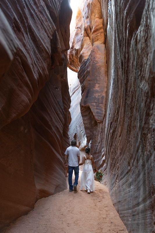 Couple in white walks hand-in-hand through a narrow red rock slot canyon with towering sandstone walls.
