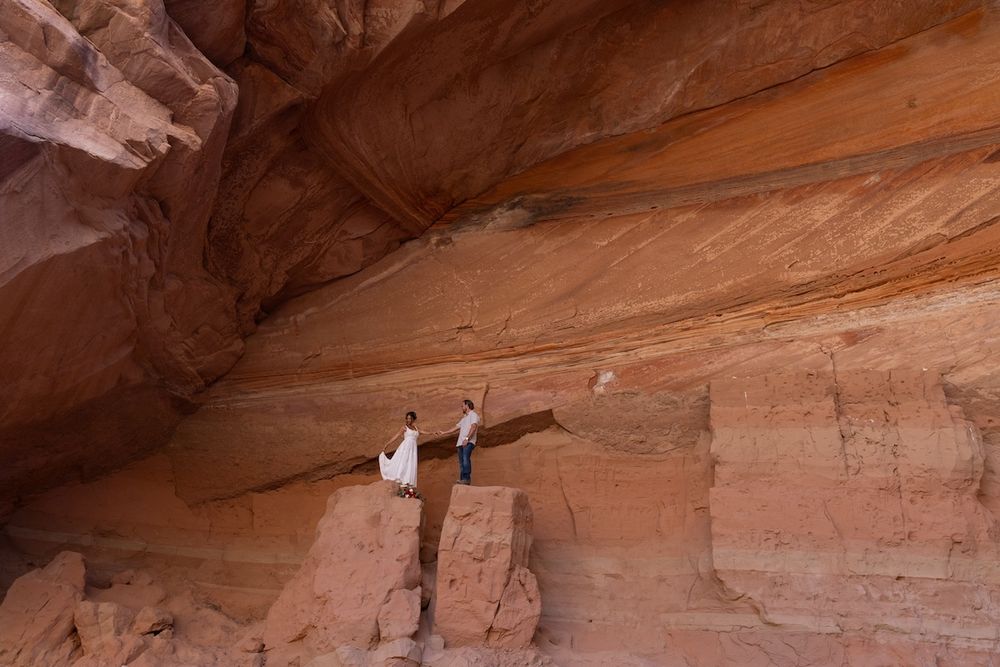 Couple in wedding attire standing on red sandstone canyon wall ledge in dramatic desert landscape.