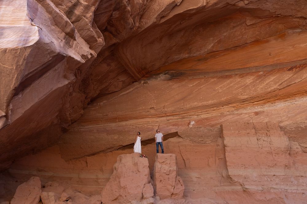 Couple in wedding attire standing beneath a massive red sandstone rock overhang in a desert canyon.