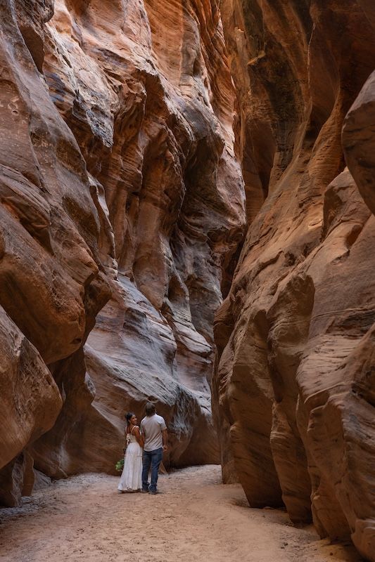 Couple in formal wear exploring a narrow red rock slot canyon with towering sandstone walls.