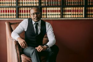 Professional man in vest and tie seated in leather chair before law library bookshelves.