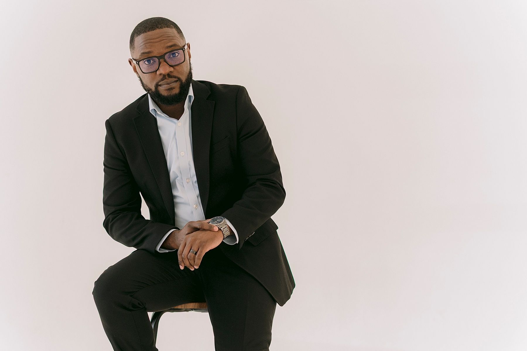Professional man in black suit and glasses sitting confidently against white background in formal portrait photo.