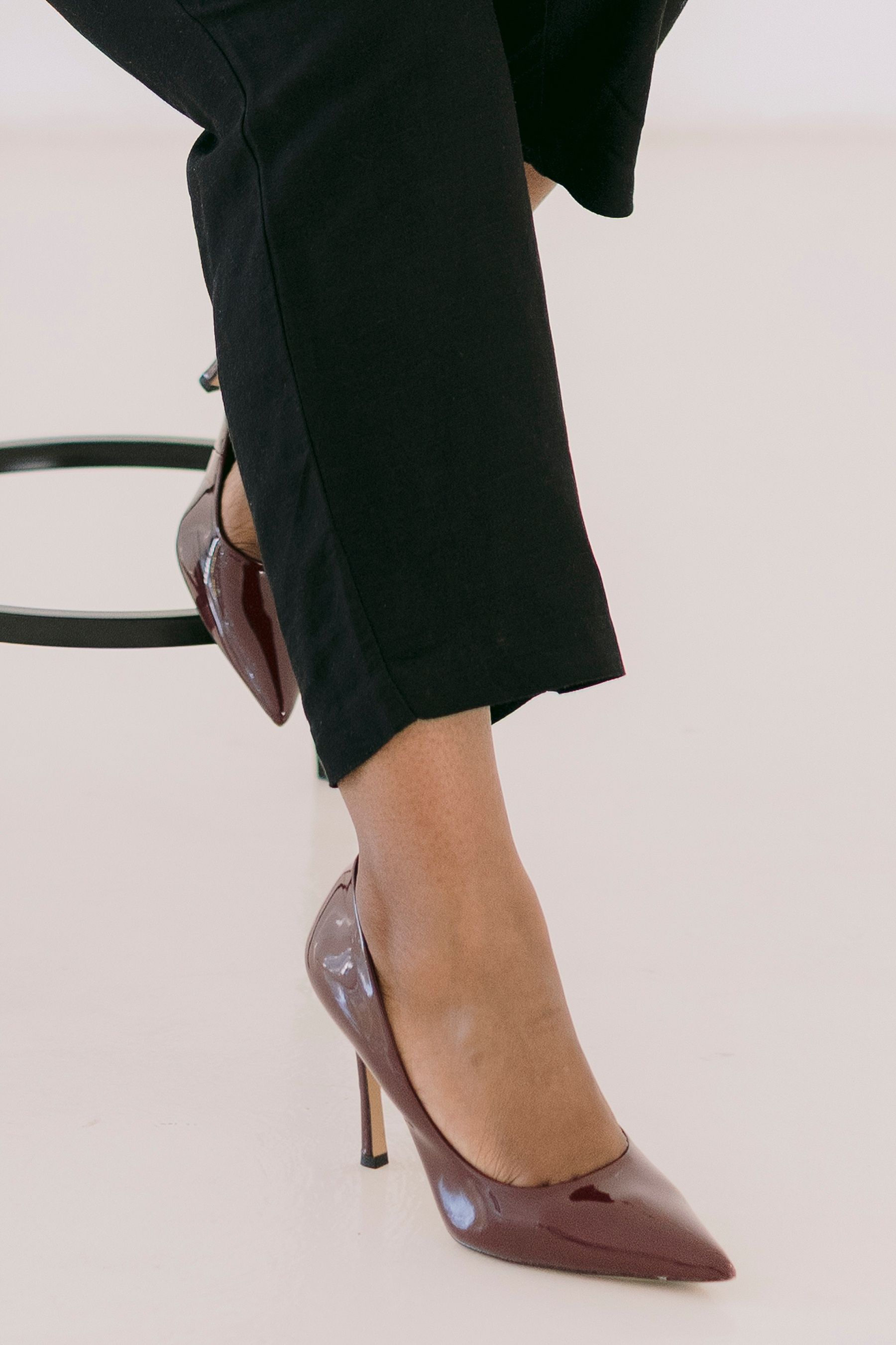 Woman wearing black cropped trousers and burgundy patent leather pointed-toe stiletto heels, seated on a stool.