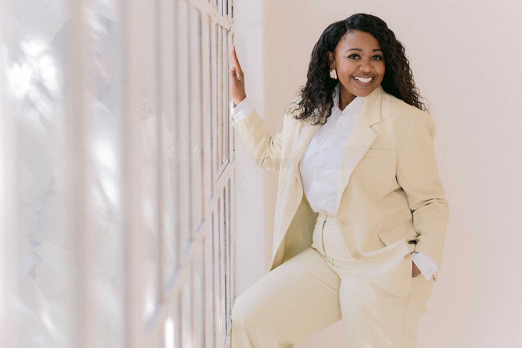 A confident woman in a cream blazer and pants smiles while leaning against a white wall in a professional pose.