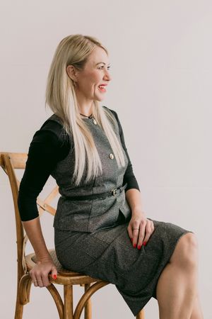 Smiling blonde woman in gray tweed dress and black top sitting on wooden chair against white background.