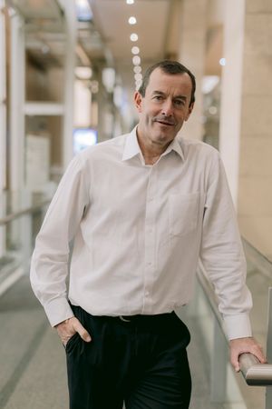 A smiling middle-aged man in a white dress shirt and black trousers stands confidently in a modern corridor.