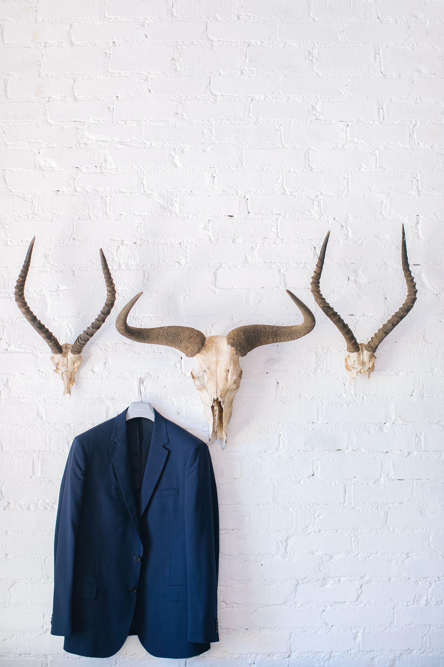 Navy blue suit jacket hanging on white brick wall beneath mounted animal skulls with horns as rustic decor.