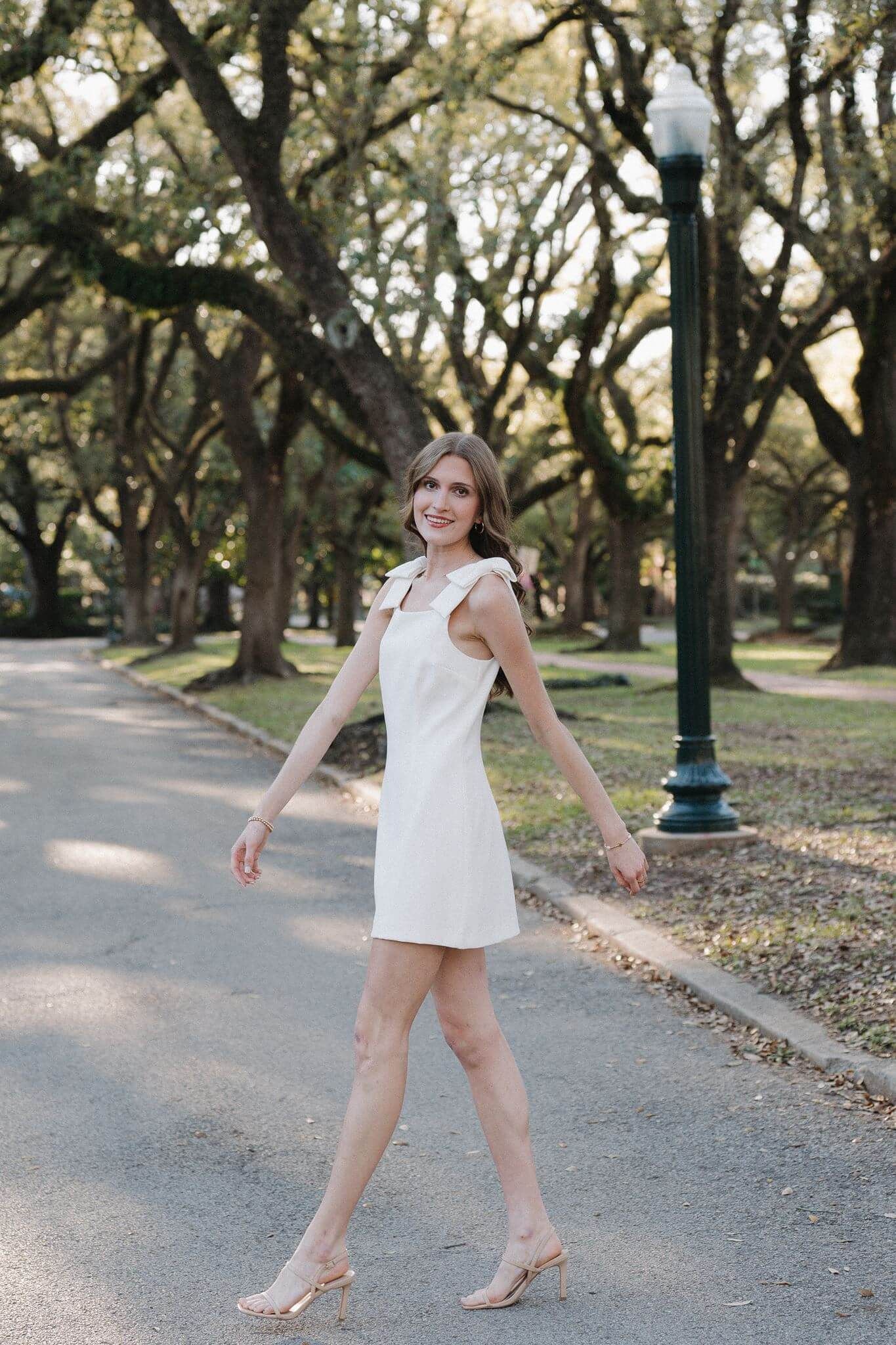 College Park High School senior girl, wearing short white dress, walking under oak tree canopy in Houston Texas.