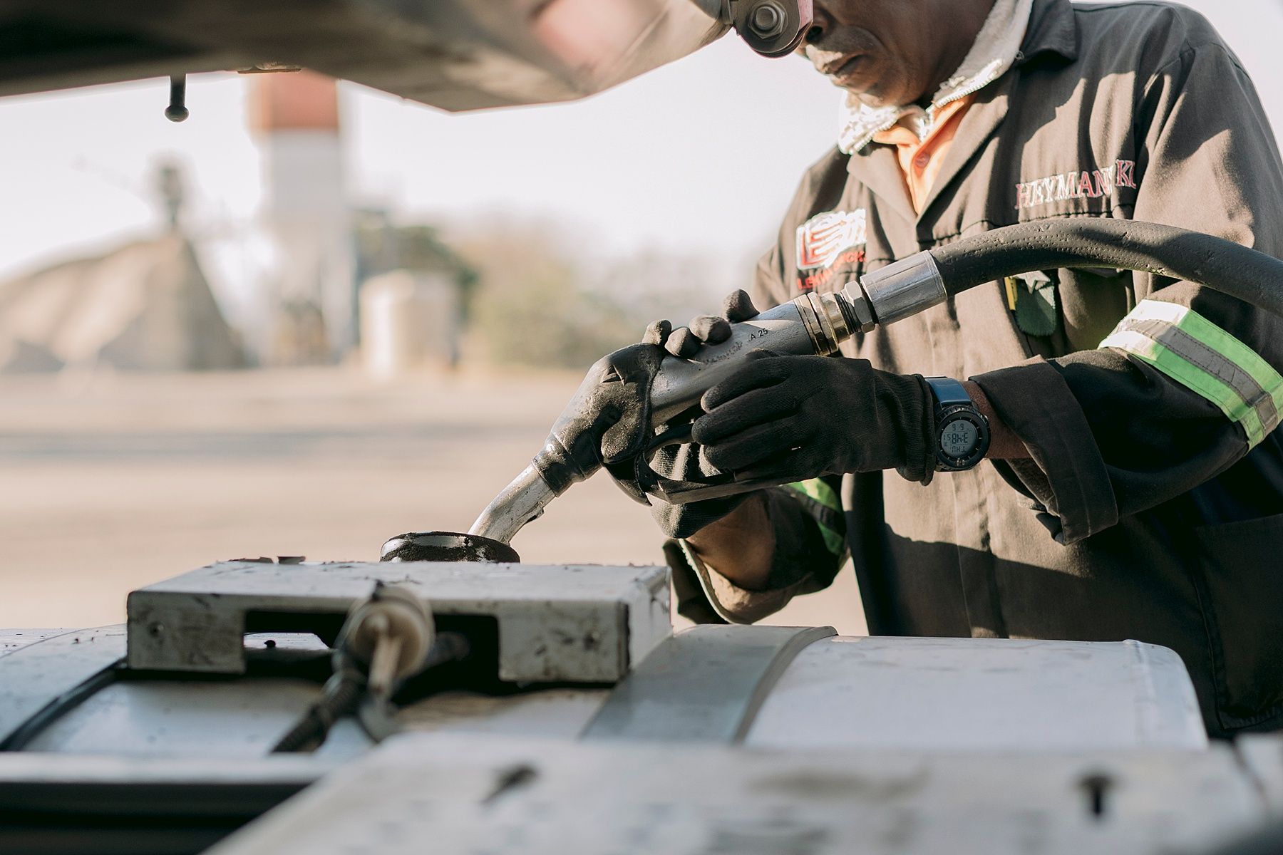 Mechanic in work uniform using a tool under the hood of a vehicle outdoors at an auto repair shop.