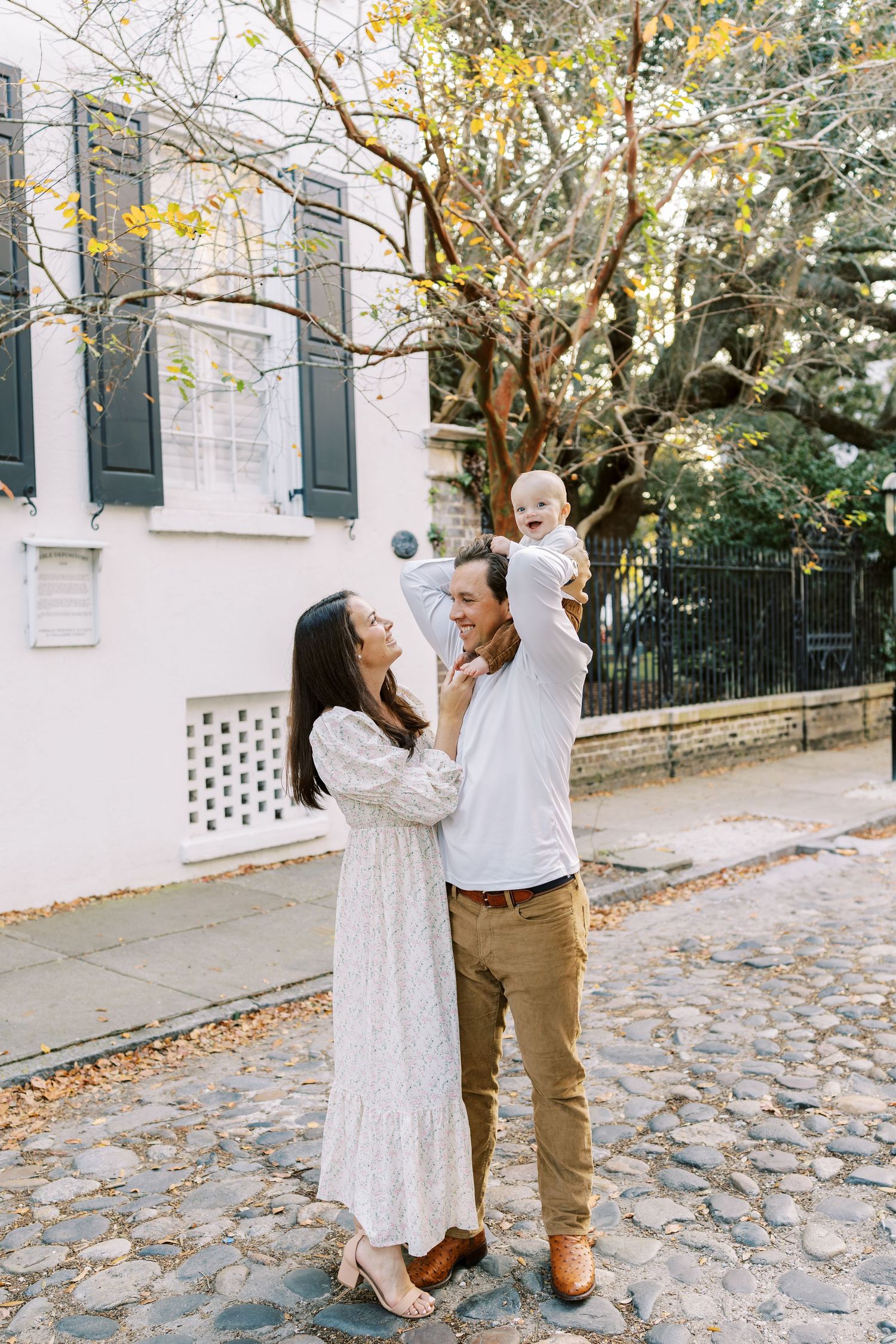 A happy young family with a baby outdoors on cobblestone street, surrounded by autumn trees and a white historic building.