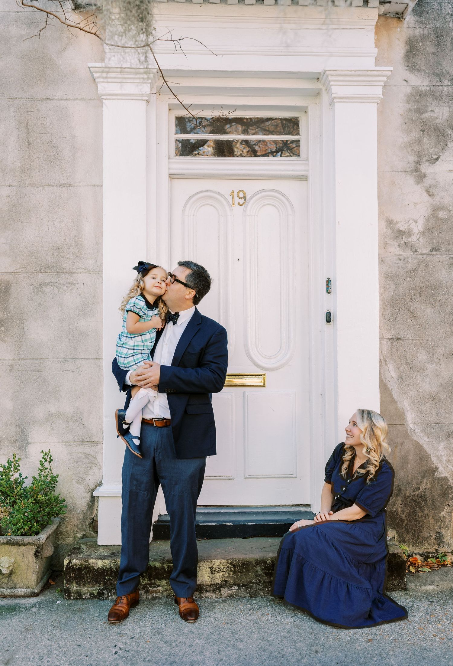 Family portrait of man holding toddler and woman in navy dress posing by white door numbered 19.