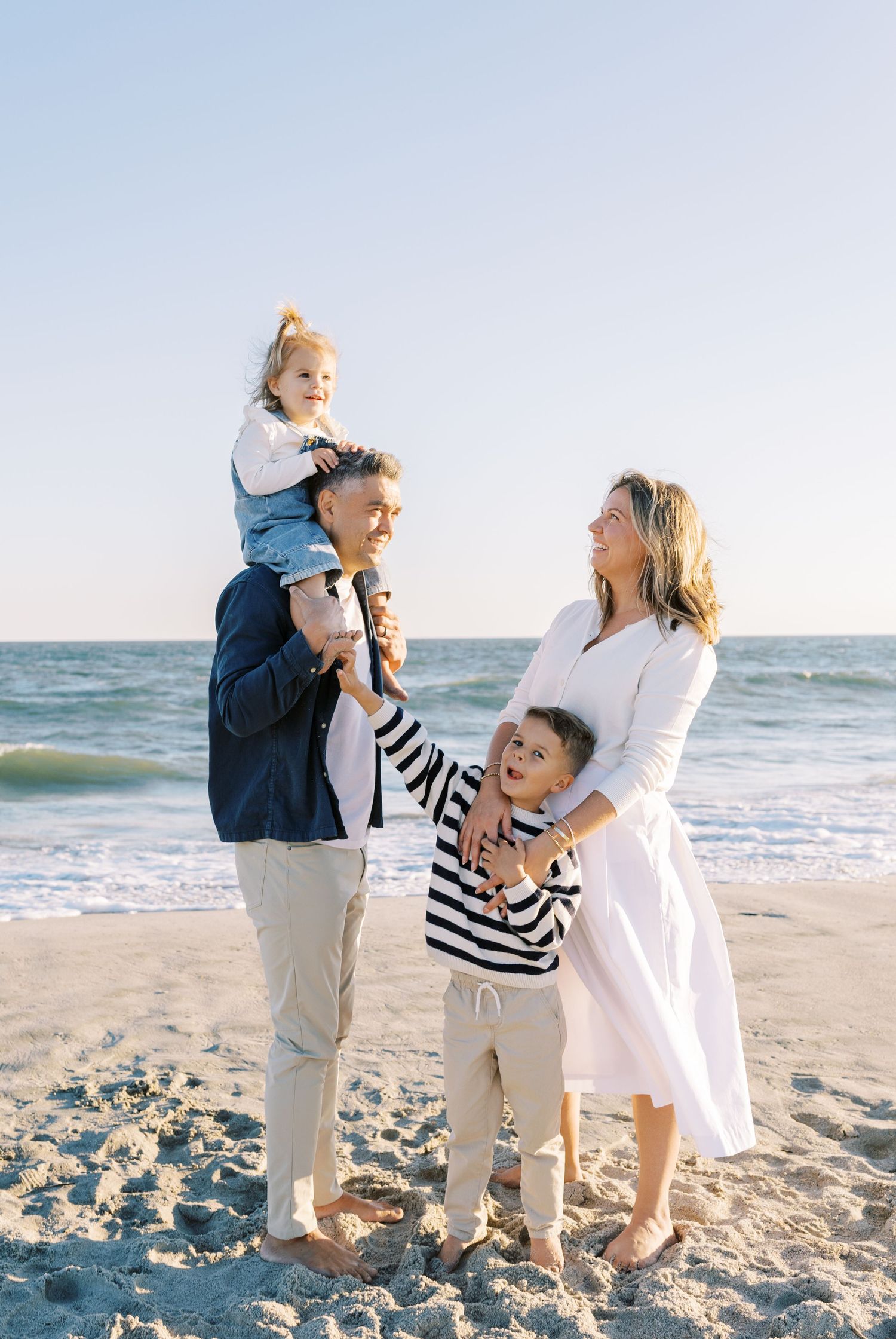 Family of four posing on sandy beach, father carrying toddler on shoulders, mother in white dress, son in striped shirt.