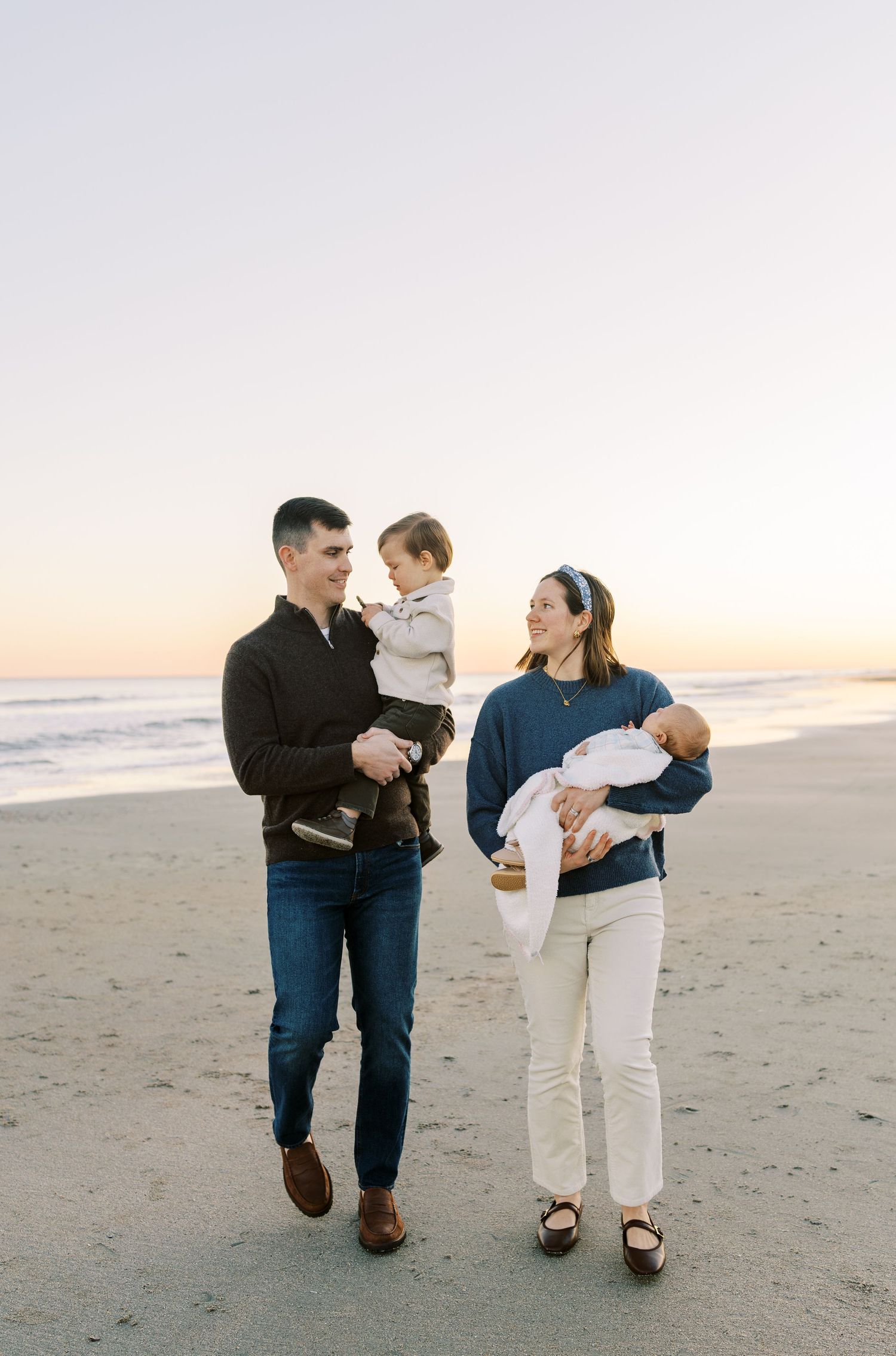 A young family walks on a beach at sunset, parents carrying two young children, dressed in casual navy and neutral tones.