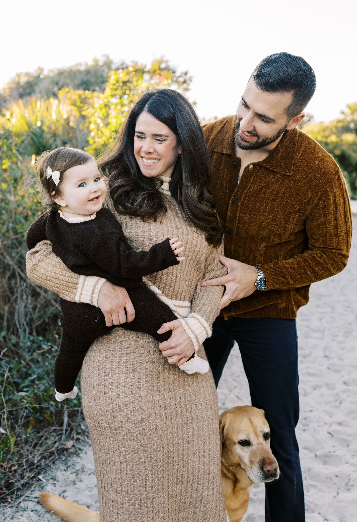 Happy family with toddler and golden dog posing outdoors on sandy path surrounded by greenery.
