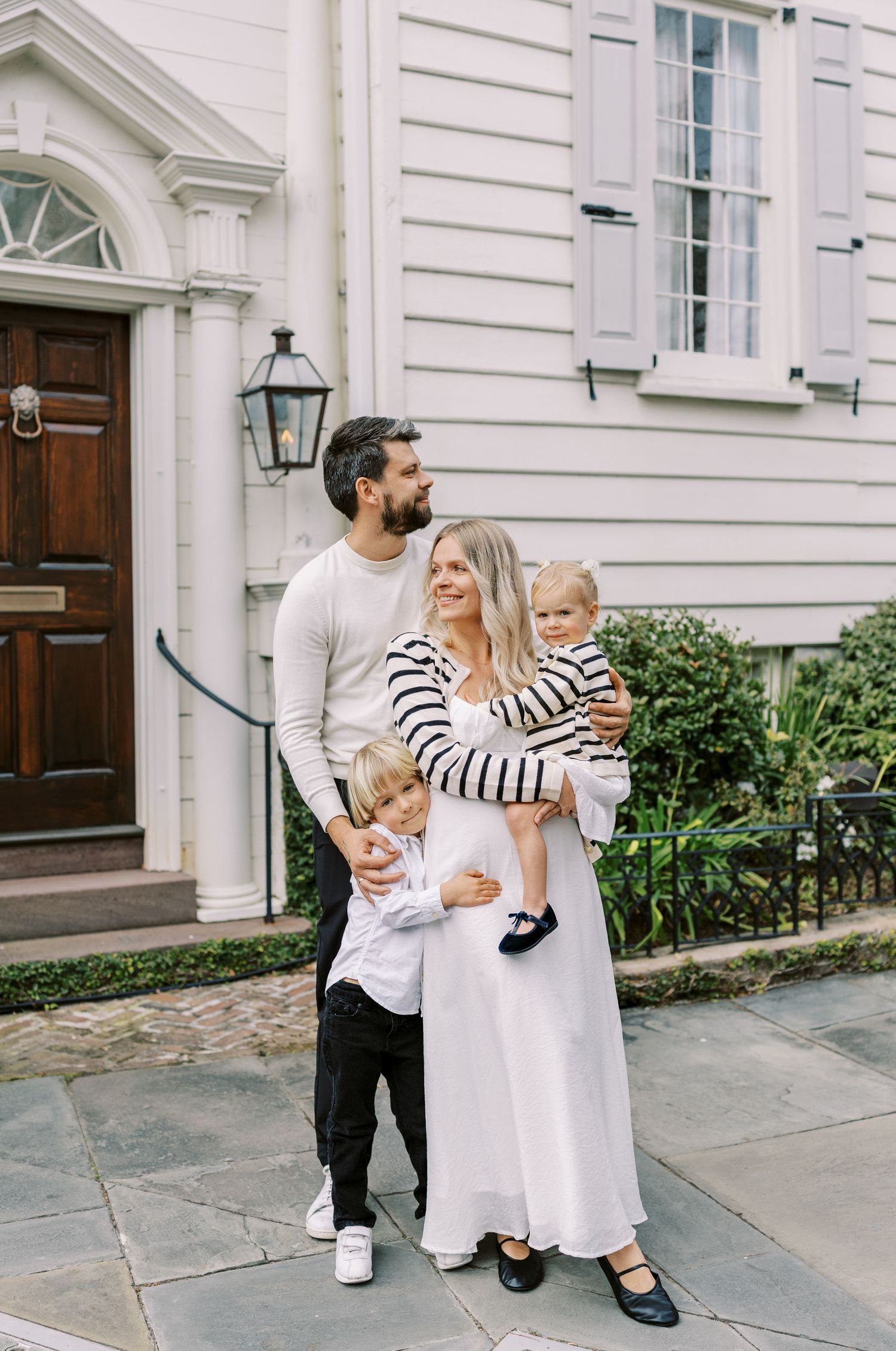 A happy family of four poses outside a white colonial home, with parents and two young children dressed in coordinating outfits.