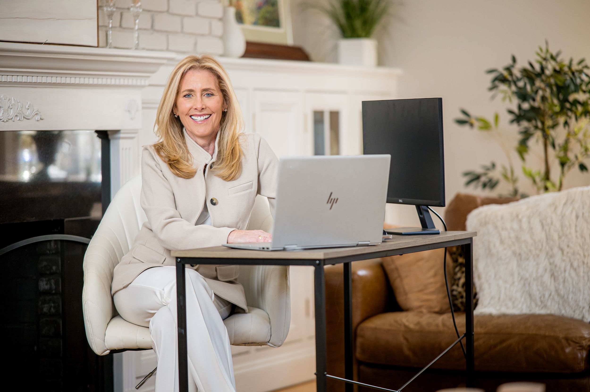 Professional woman smiling at desk with HP laptop and monitor in elegant home office near fireplace.