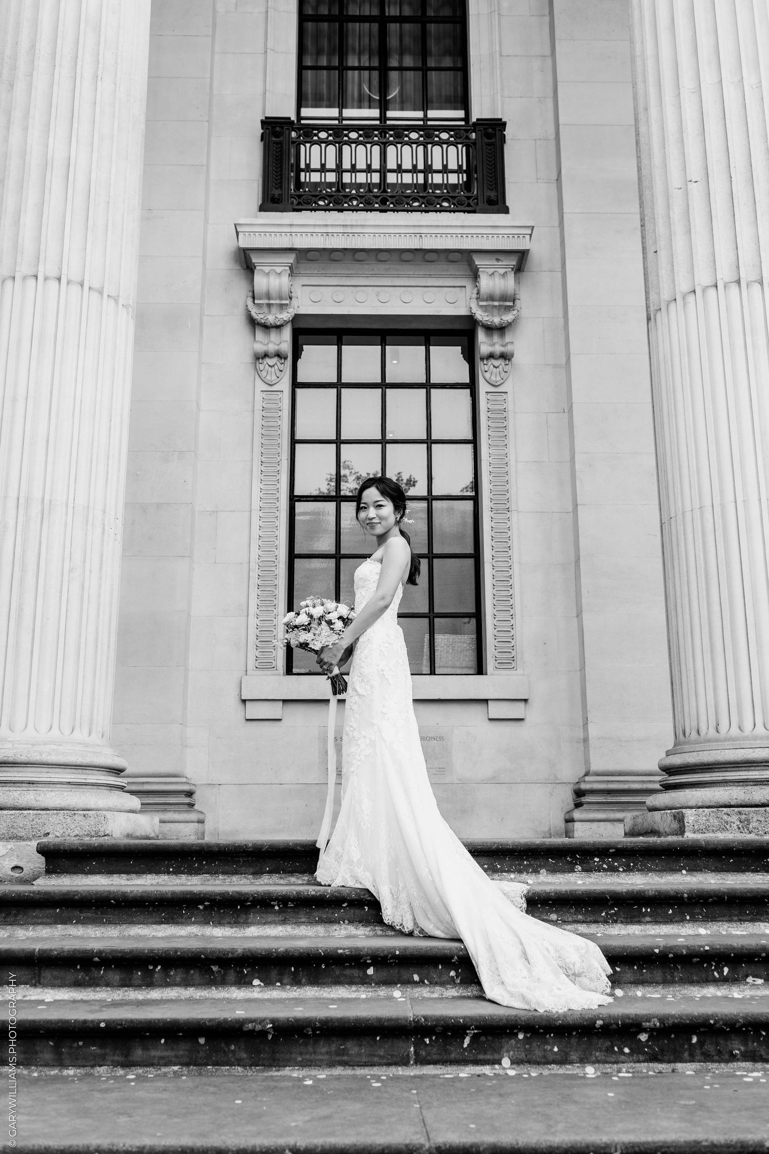 Black and white photo of a bride in a white gown holding a bouquet on steps of a grand building with columns.