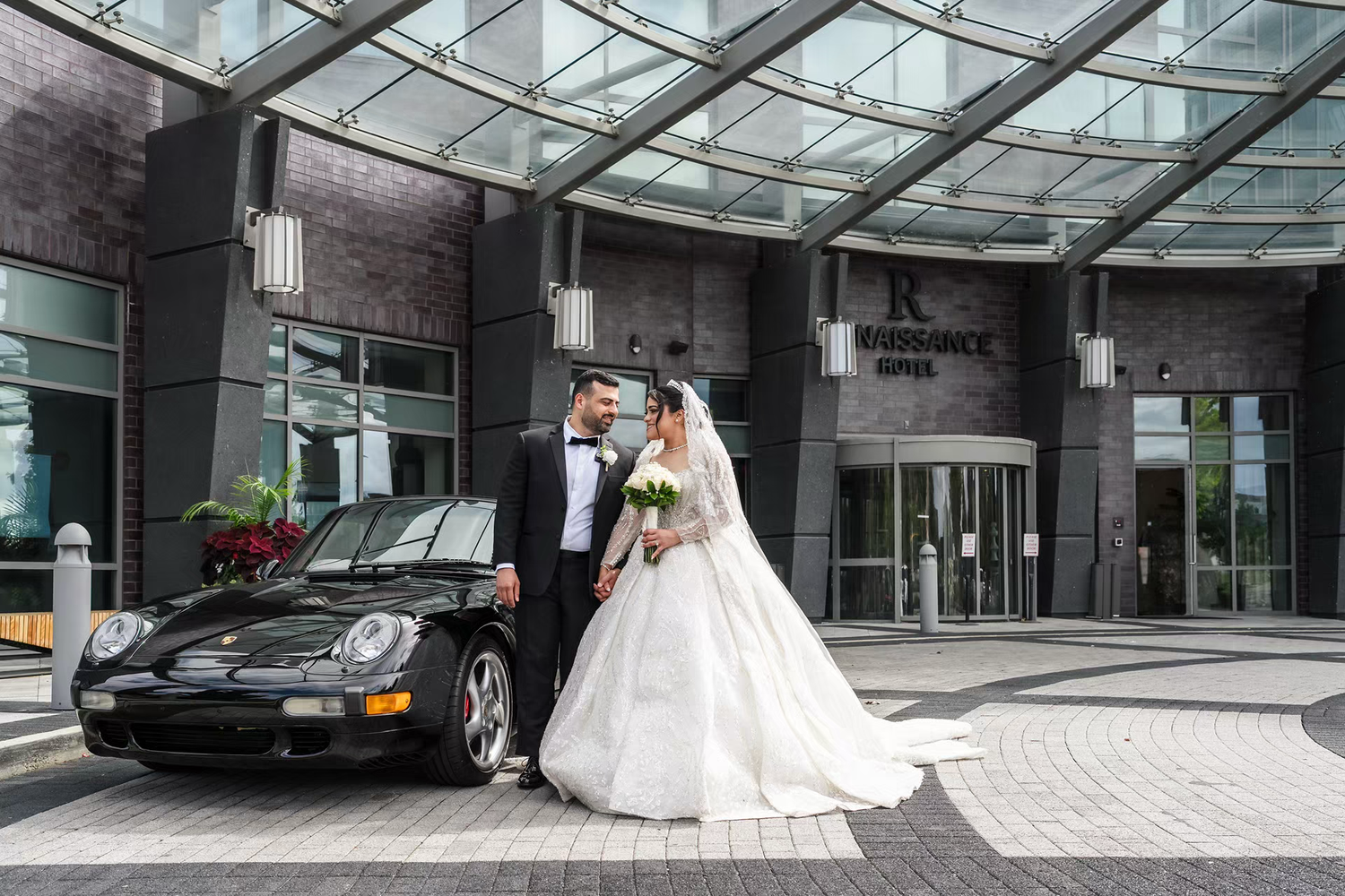 Bride and groom posing in front of a black Porsche at a renaissance hotel entrance on their wedding day.