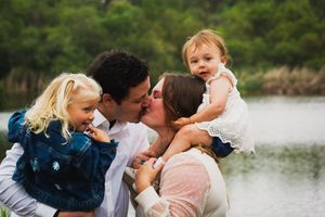 A happy couple kissing while holding their two young toddler daughters near a scenic lake outdoors. Vista, CA