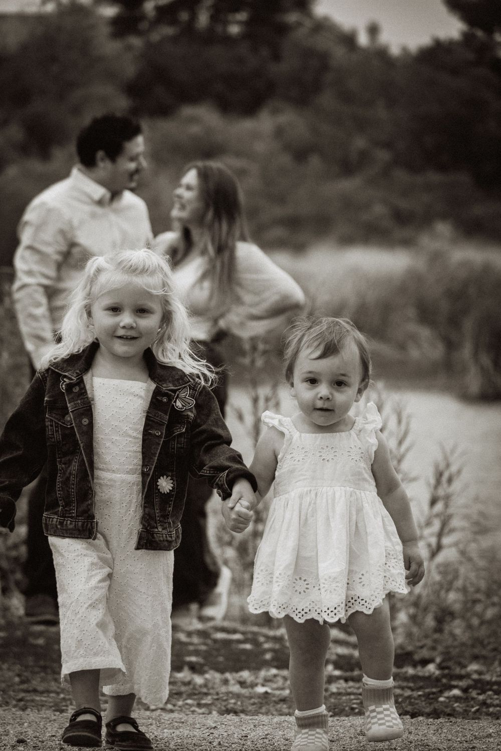 Black and white family photo of two young girls in white dresses holding hands while parents share a tender moment behind them. Vista, CA