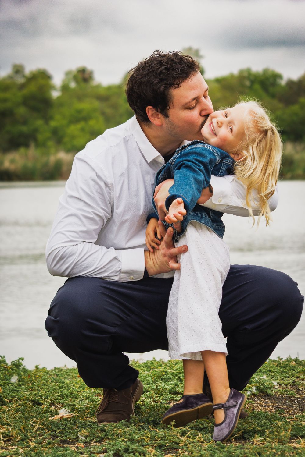 Father crouching by a lake, kissing his smiling blonde toddler daughter outdoors near lush greenery. Vista, CA